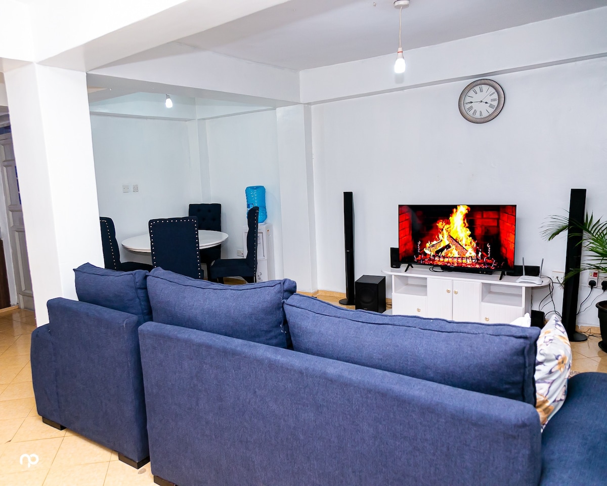 A spacious living area is shown, featuring a blue sectional sofa facing a modern television with a digital fireplace display. A round dining table with upholstered chairs is visible in the background, alongside a wall clock, creating a welcoming space for relaxation and gatherings.