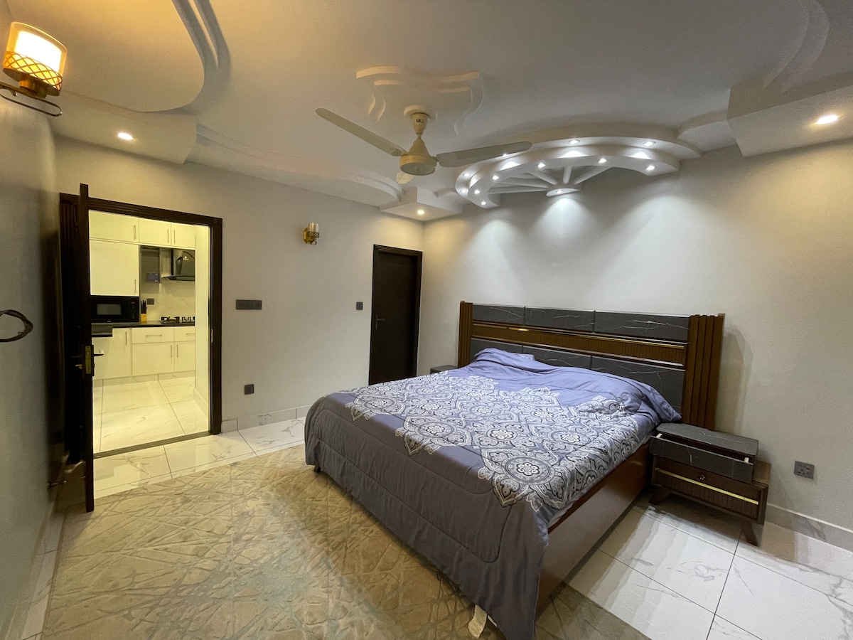 A spacious bedroom features a large bed with a decorative comforter. Soft lighting is provided by built-in ceiling fixtures, and a contemporary ceiling fan is visible. A door leads to a kitchenette area, enhancing the room's functionality. A plush area rug is placed on the tiled floor.