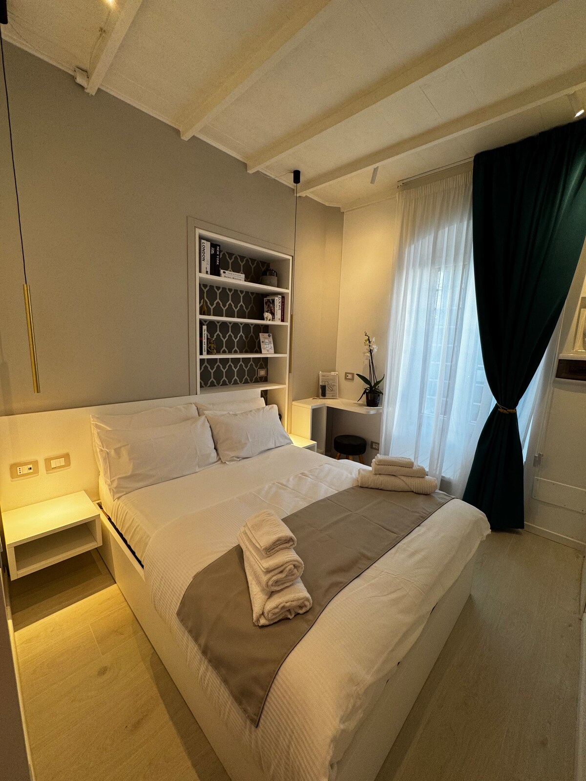 A compact sleeping area features a spacious bed with neutral bedding and decorative pillows. A bedside table and a small shelf are visible beside the bed, while a tall window is draped with a green curtain. Natural light brightens the room, creating a serene ambiance.