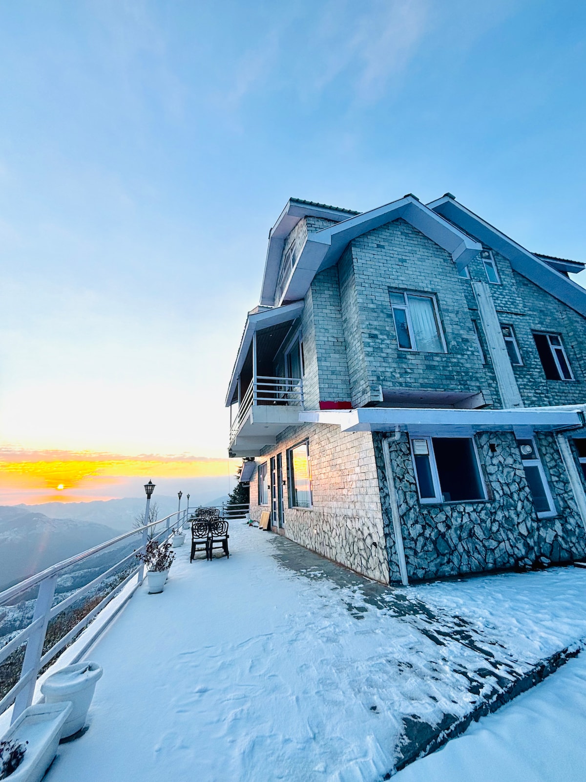 The building is situated on a snowy slope, showcasing a mixture of stone and contemporary architecture. The pathway is lined with potted plants, and outdoor seating is available. A sunset casts soft colors across the sky, enhancing the serene mountain backdrop.