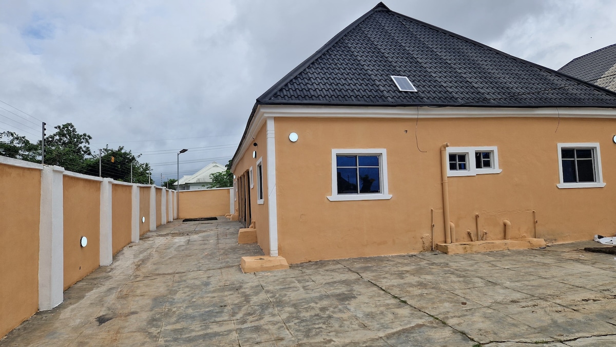 The exterior of the building is presented, featuring a single-story structure with a distinctive dark tiled roof. The walls are a warm beige, and several windows allow natural light to enter. A spacious, paved area is visible in front, complemented by a secure perimeter wall.