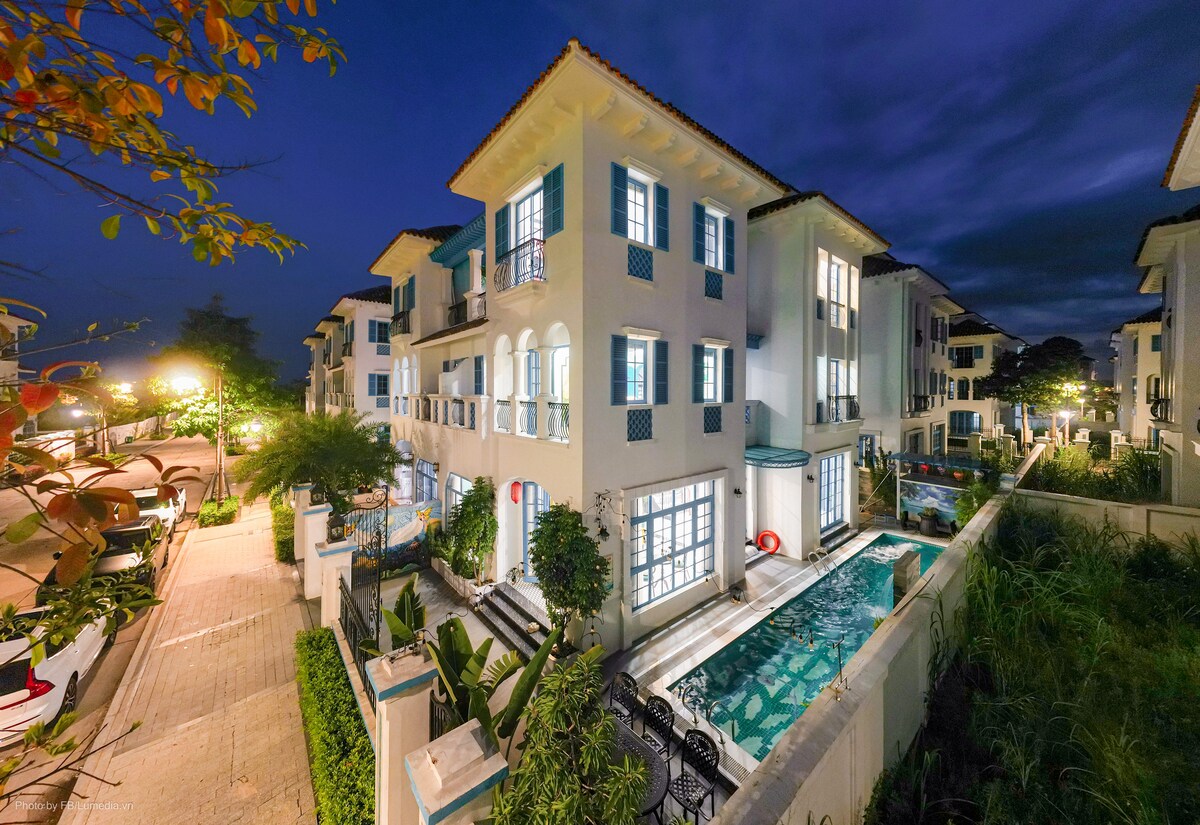 The villa's exterior is showcased at dusk, featuring a combination of white walls and blue accents. Large windows reflect soft lighting, and lush greenery surrounds the property. A tranquil pool can be seen, enhancing the peaceful ambiance of the setting.