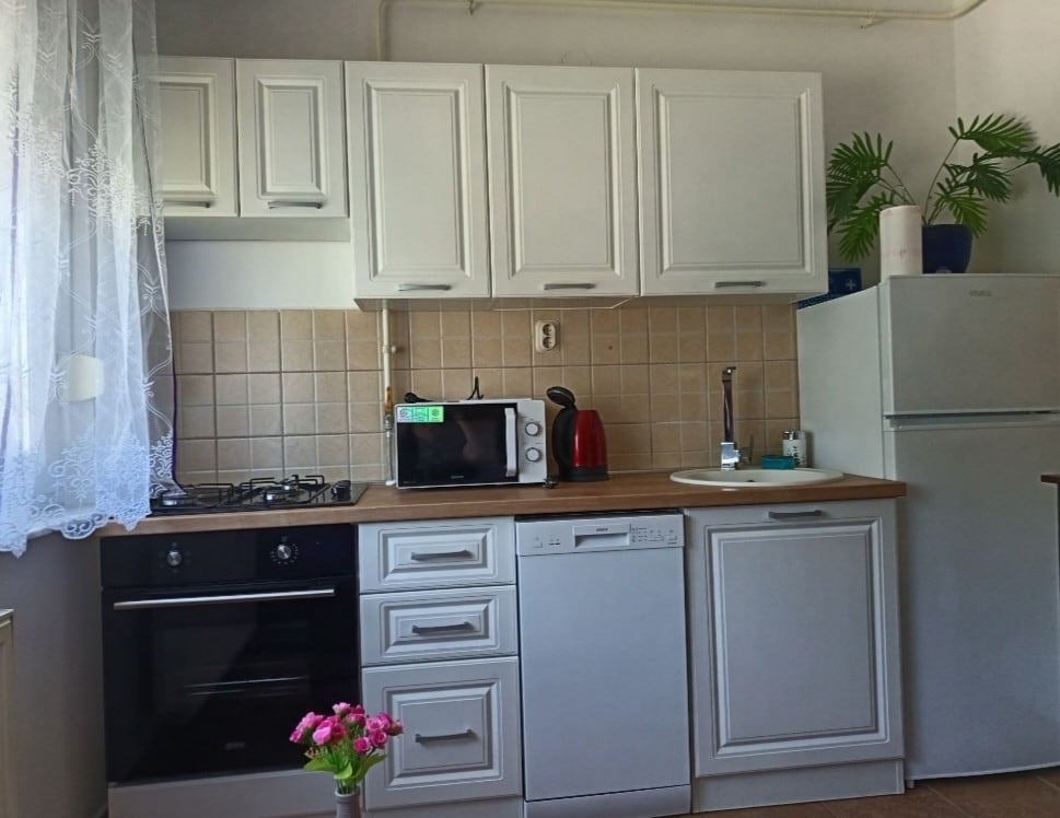A well-equipped kitchen features modern cabinetry in white, complemented by a tiled backsplash. Essential appliances, including an oven, microwave, kettle, and refrigerator, are visible. A small bouquet of flowers adds a touch of warmth, while a window dressed in lace lets in natural light.