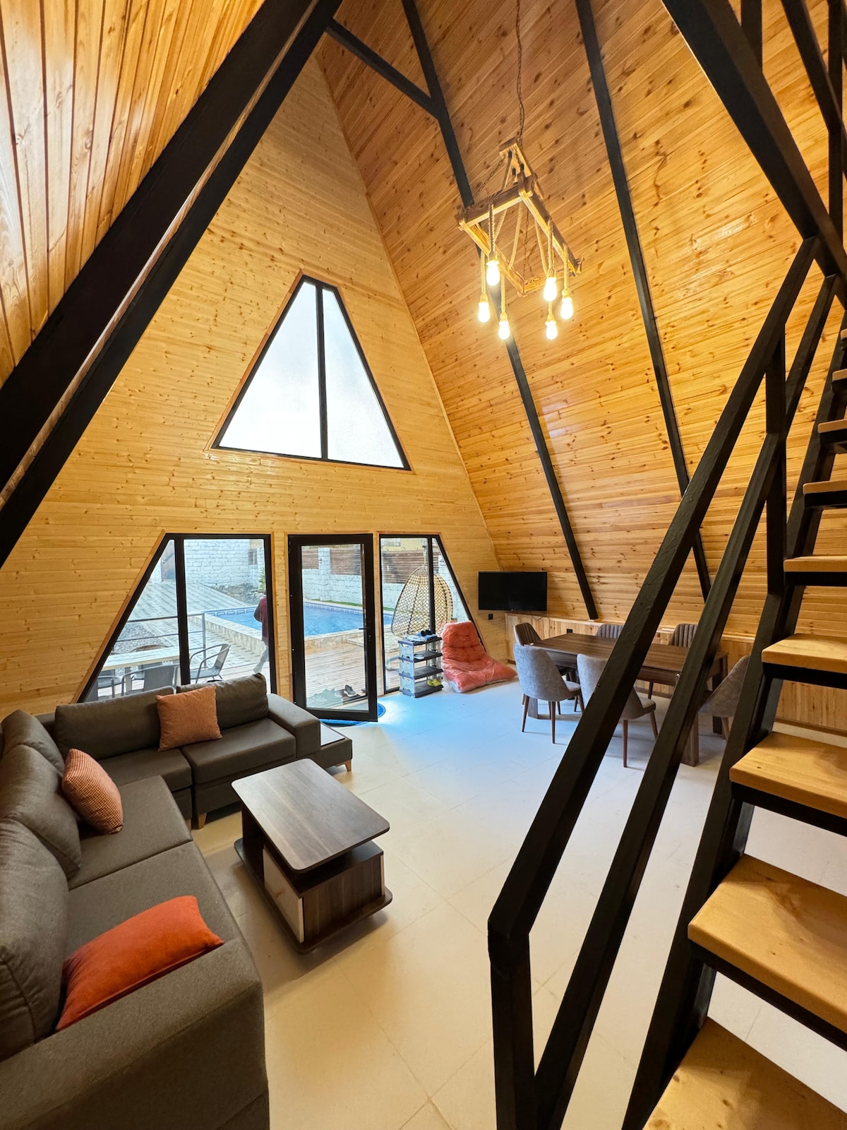 The interior features a high triangular ceiling with wooden paneling, complemented by large windows allowing natural light to fill the space. A cozy seating area is arranged around a coffee table, and a dining area is positioned nearby, providing a comfortable gathering spot.
