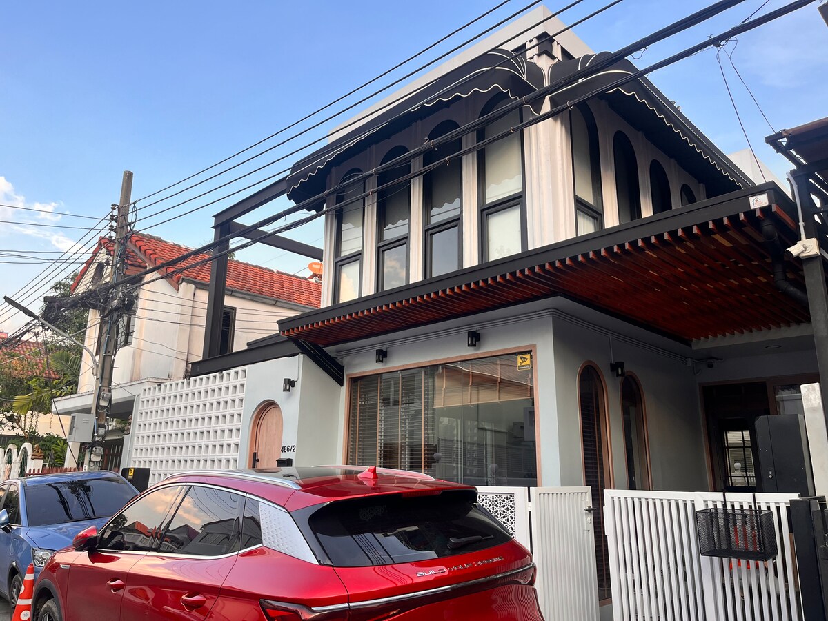 The exterior of a stylish house is displayed, showcasing a combination of modern and traditional architectural elements. A red car is parked in front, with the building featuring large windows and a distinctive balcony design. Clear blue skies provide a bright backdrop.