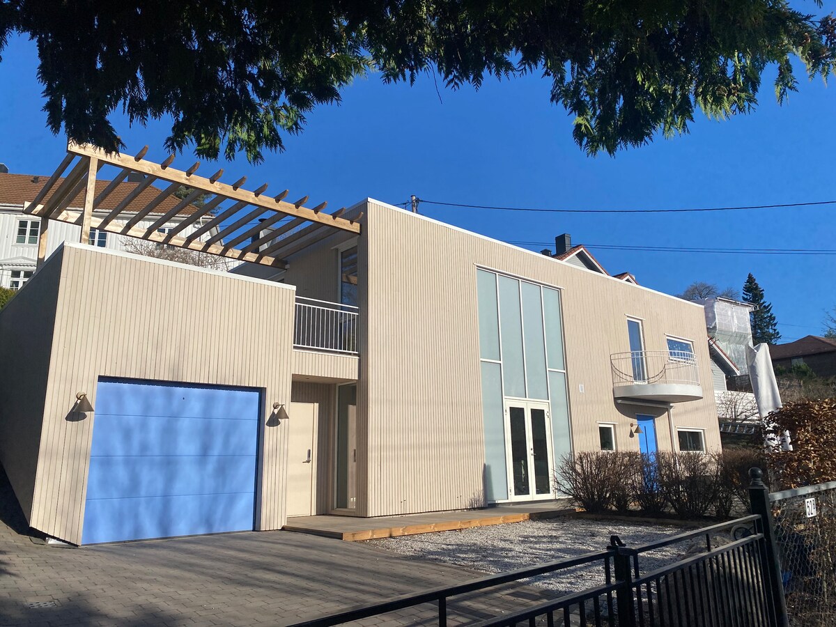 The modern house is presented with a light-colored exterior, featuring multiple large windows for natural light. A blue garage door adds a pop of color. A wooden deck extends from the entrance, and the upper level has an overhang supported by beams, enhancing the architectural interest.