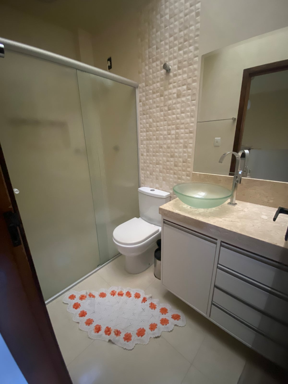 A compact bathroom features a glass shower enclosure and a modern toilet. A bowl sink is positioned on a light-colored countertop with storage cabinets underneath. A patterned rug sits on the tiled floor, adding a touch of color to the space.