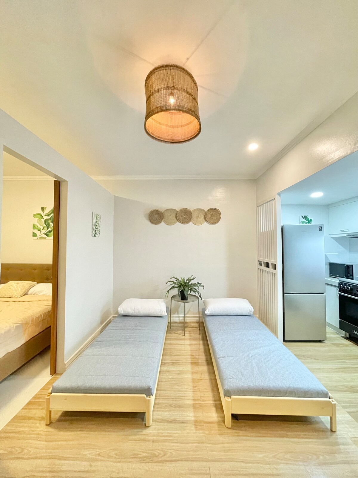 The living area features two stackable beds positioned side by side, with minimal bedding and gray covers. A small potted plant adds a touch of greenery. The adjacent kitchen is visible with appliances, along with a soft pendant light illuminating the space.