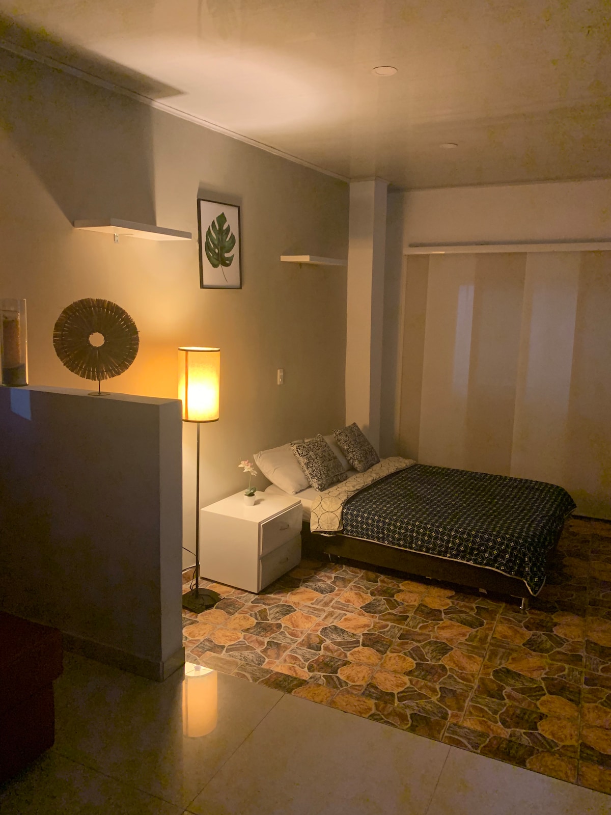 A sleeping area is featured with a spacious bed adorned in patterned bedding. Light is emitted from a standing lamp, illuminating the room's neutral walls. A small nightstand holds a decorative item, while light filters through translucent curtains, enhancing the serene environment.