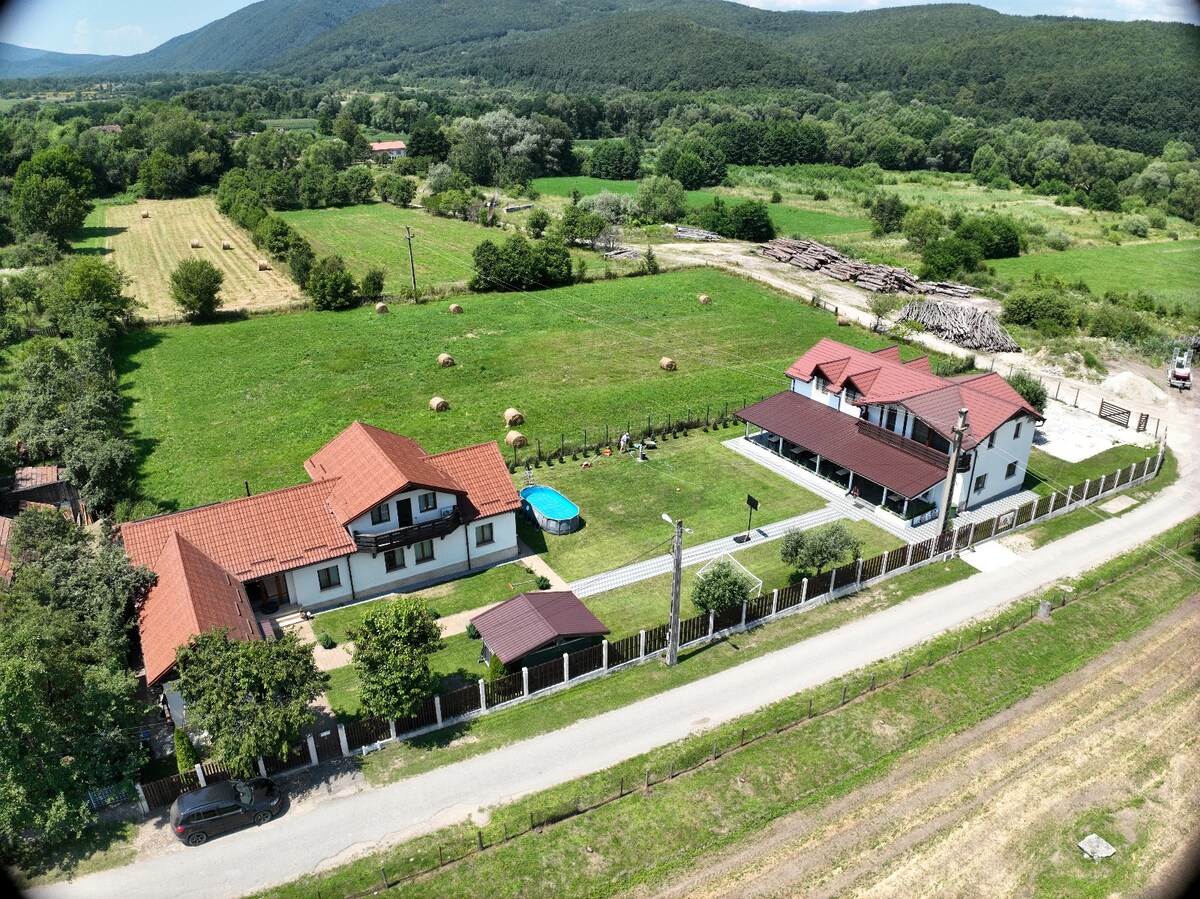 An aerial view of the property reveals multiple buildings with distinctive red roofs set against lush green fields. A swimming pool is visible surrounded by landscaped lawns, while trees and hills create a serene backdrop. Paths connect the structures, enhancing the overall spaciousness.