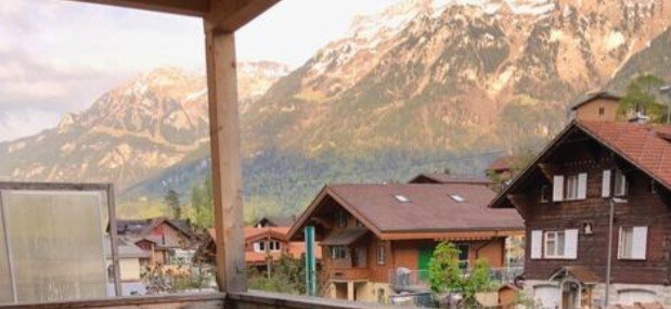 A spacious terrace offers a stunning view of the surrounding mountains and village. Nearby houses showcase a blend of traditional and modern architectural styles. The tranquil atmosphere is enhanced by the distant peaks and lush greenery, providing an inviting space for relaxation.