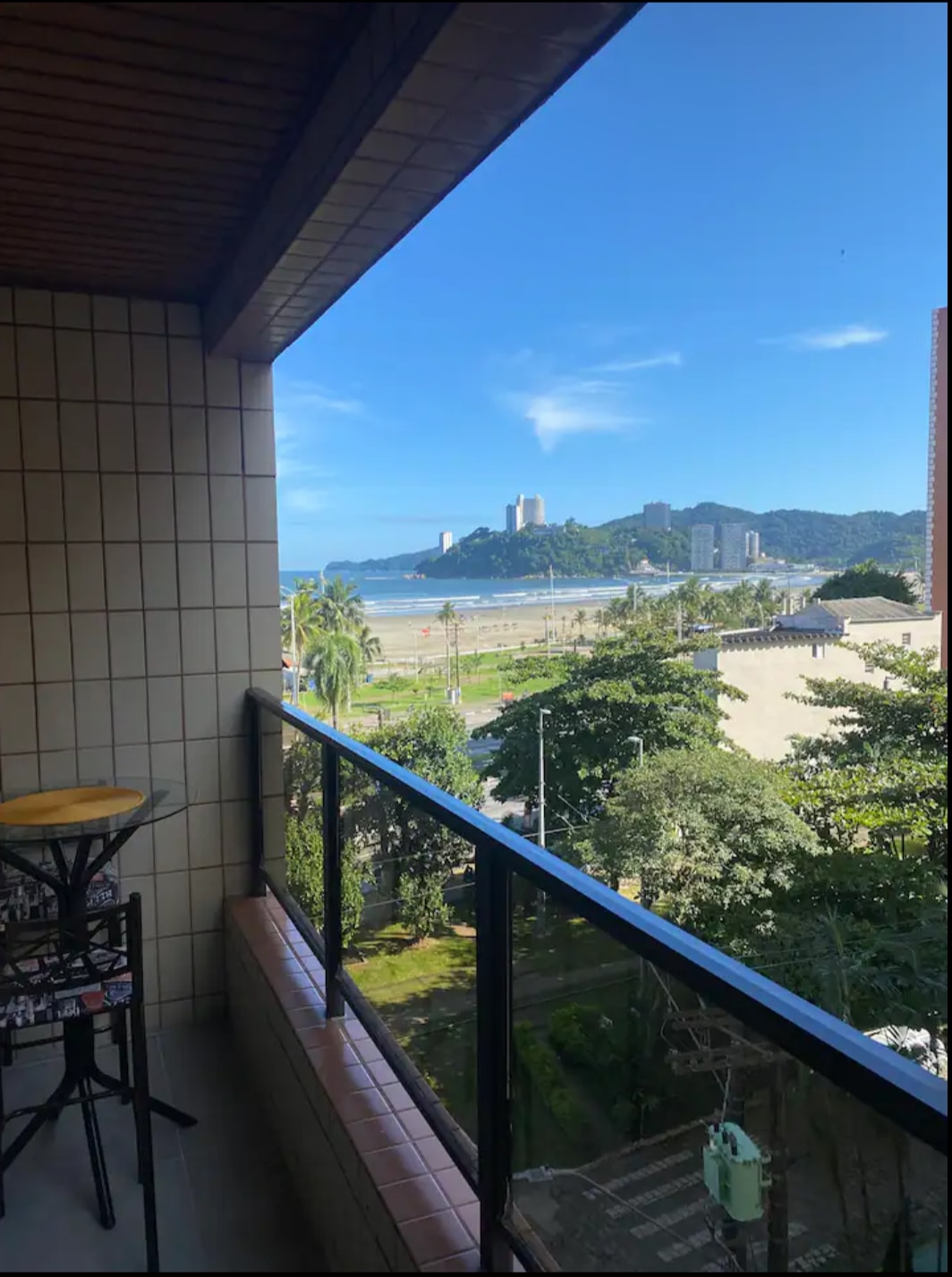 A balcony offers a view of the beach and coastline, framed by lush greenery and palm trees. The distant skyline is visible with buildings set against a bright blue sky, enhancing the outdoor ambiance.