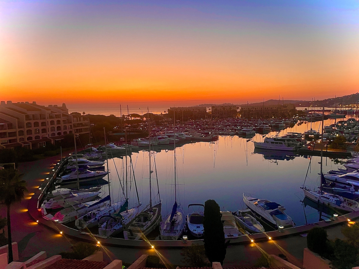 A panoramic view of a marina at sunset is captured, showcasing numerous boats docked in calm waters. The sky transitions from orange to purple, reflecting on the water's surface. Soft lights illuminate the marina's pathways, creating a serene ambiance.