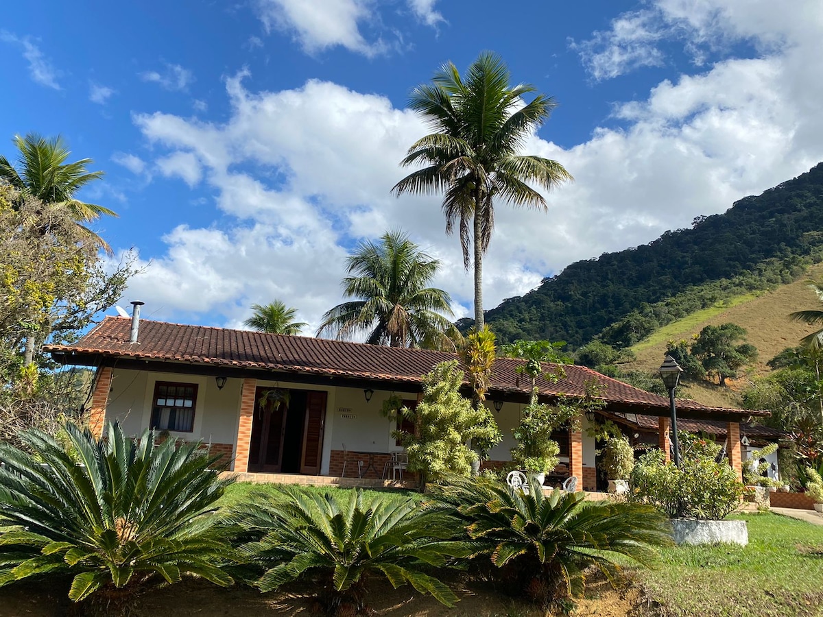 A spacious house is set against a backdrop of hills and lush greenery. Palm trees and ornamental plants are planted in the foreground. The roof features brown tiles, and large windows allow natural light to illuminate the exterior.