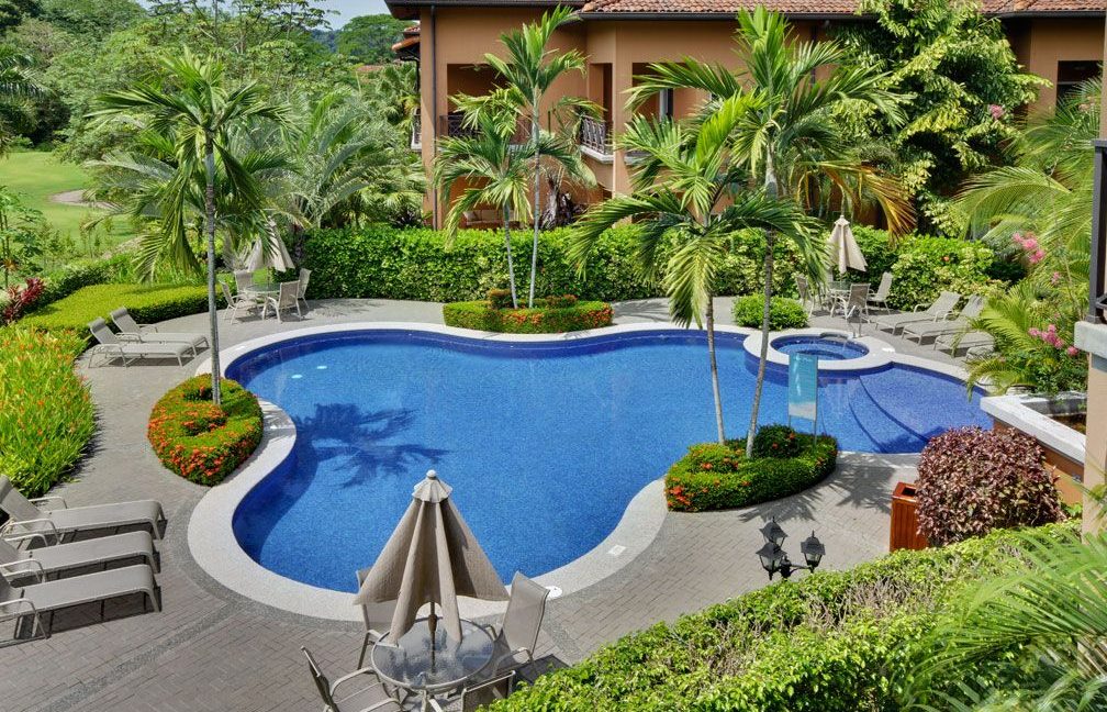 A serene swimming pool is surrounded by colorful tropical landscaping, featuring lush greenery and vibrant flowers. Multiple lounge chairs are positioned around the pool, along with umbrellas providing shade. The inviting water reflects the blue sky, contributing to the peaceful atmosphere.