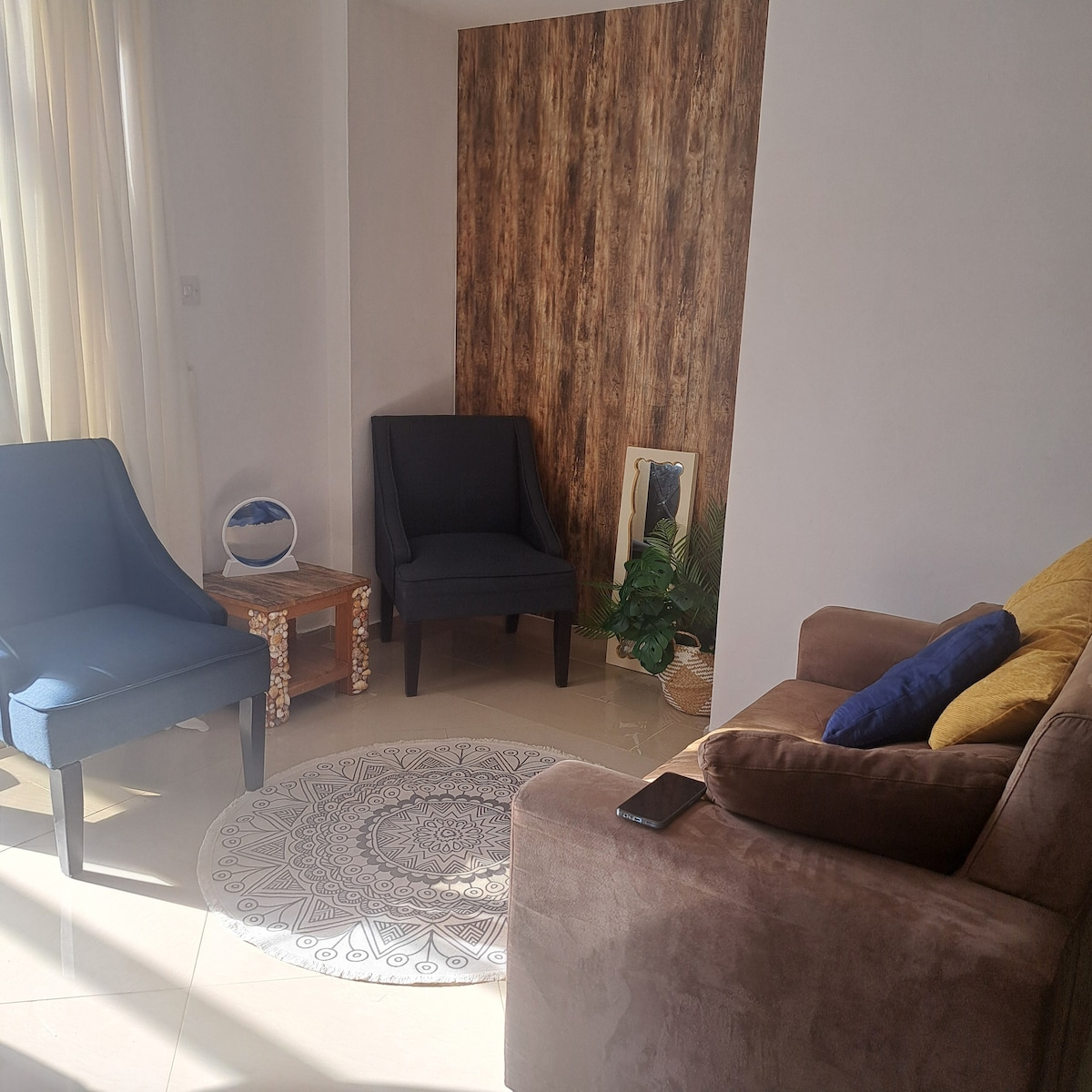 A cozy seating area features two blue chairs and a brown sofa, complemented by a decorative rug with intricate patterns. Natural light filters in through sheer curtains, creating a warm ambiance. A potted plant and a small table add to the inviting space.