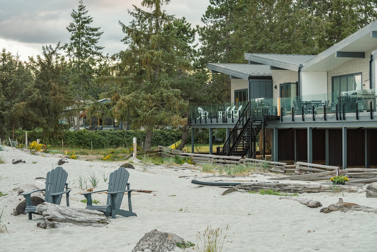 Adirondack chairs are placed on soft white sand, facing the ocean. In the background, the beach house rises on stilts with a spacious outdoor deck. Glass railings provide an unobstructed view of the surrounding natural landscape, framed by trees and coastal vegetation.