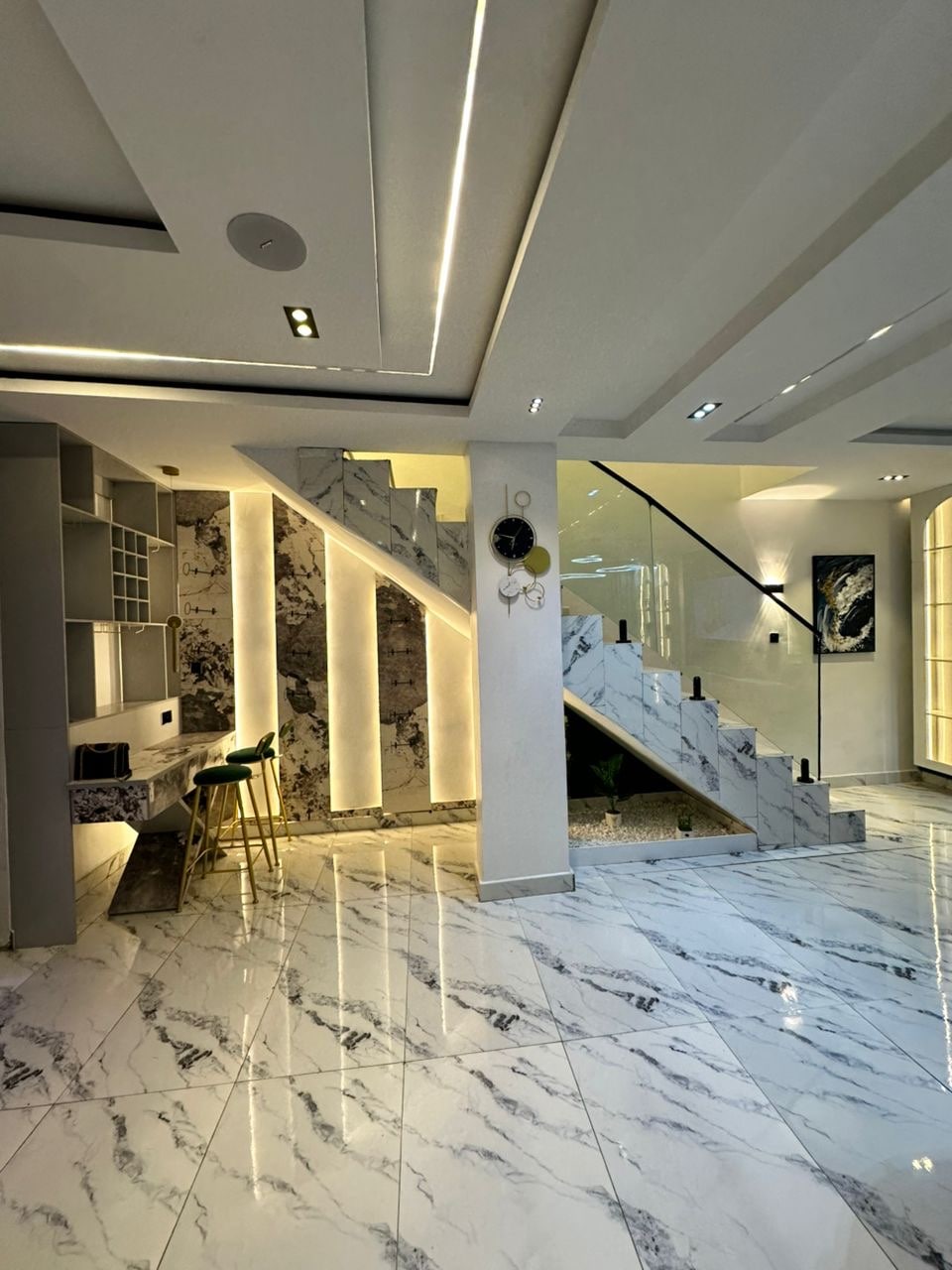 A spacious entryway is highlighted by a sleek marble floor, featuring elegant lighting along the staircase. The walls showcase a modern design with light accents, while decorative elements are positioned beside the stairs, enhancing the contemporary aesthetic.