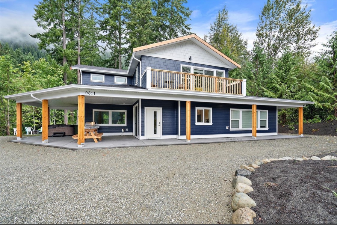 The spacious exterior of the lake house is framed by lush greenery and towering trees. A welcoming front porch is furnished with a seating area. The two-story structure features a blend of blue siding and natural wood accents, with large windows allowing for ample natural light.