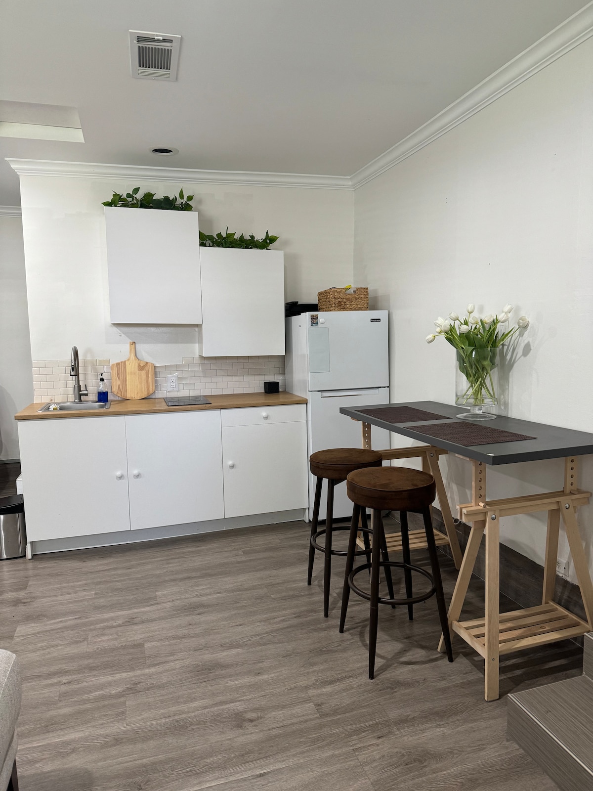 A modern kitchen area features sleek white cabinetry and a light wood countertop. A refrigerator stands beside the cabinetry. Two round bar stools are situated at a compact dining table, accented by a vase of fresh flowers, contributing to a clean and functional space.