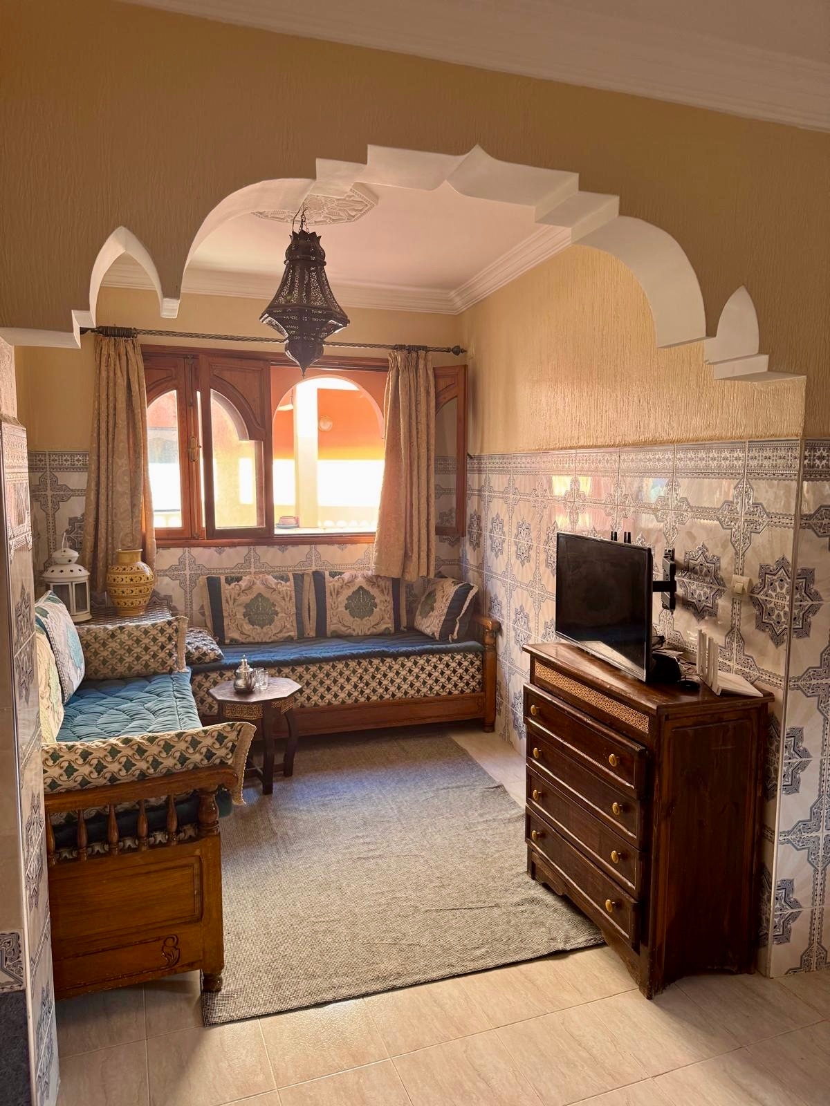 The living area features traditional Moroccan design elements, including decorative tile work and arched doorways. Comfortable seating arrangements are positioned around a small table, with natural light illuminating the space through the large windows adorned with light curtains.