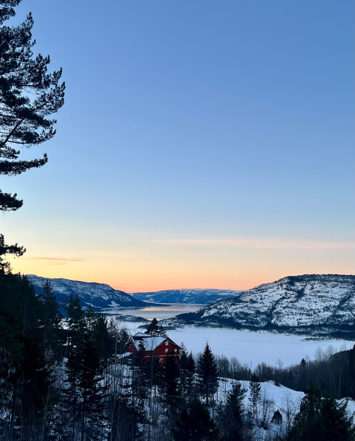 A serene twilight landscape captures the view over Nisser fjord, with snow-covered mountains framing the horizon. A traditional cabin nestled among trees is visible in the foreground, highlighting the natural beauty of the surrounding area.