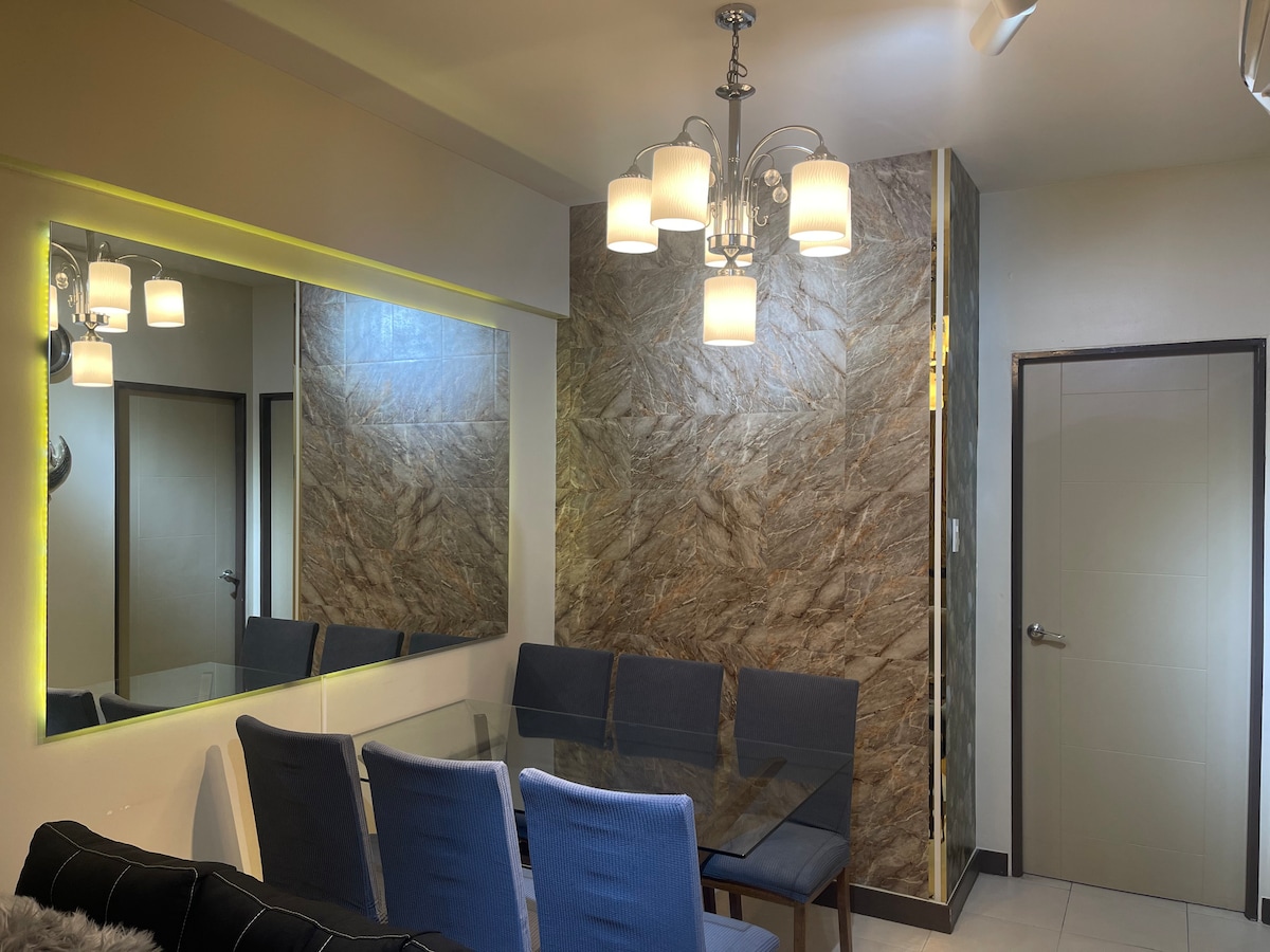 A dining area features a glass table surrounded by six blue upholstered chairs, reflecting a modern aesthetic. A wall-mounted mirror enhances the space, while a stylish chandelier illuminates the room. Textured stone wall accents add depth to the inviting atmosphere.