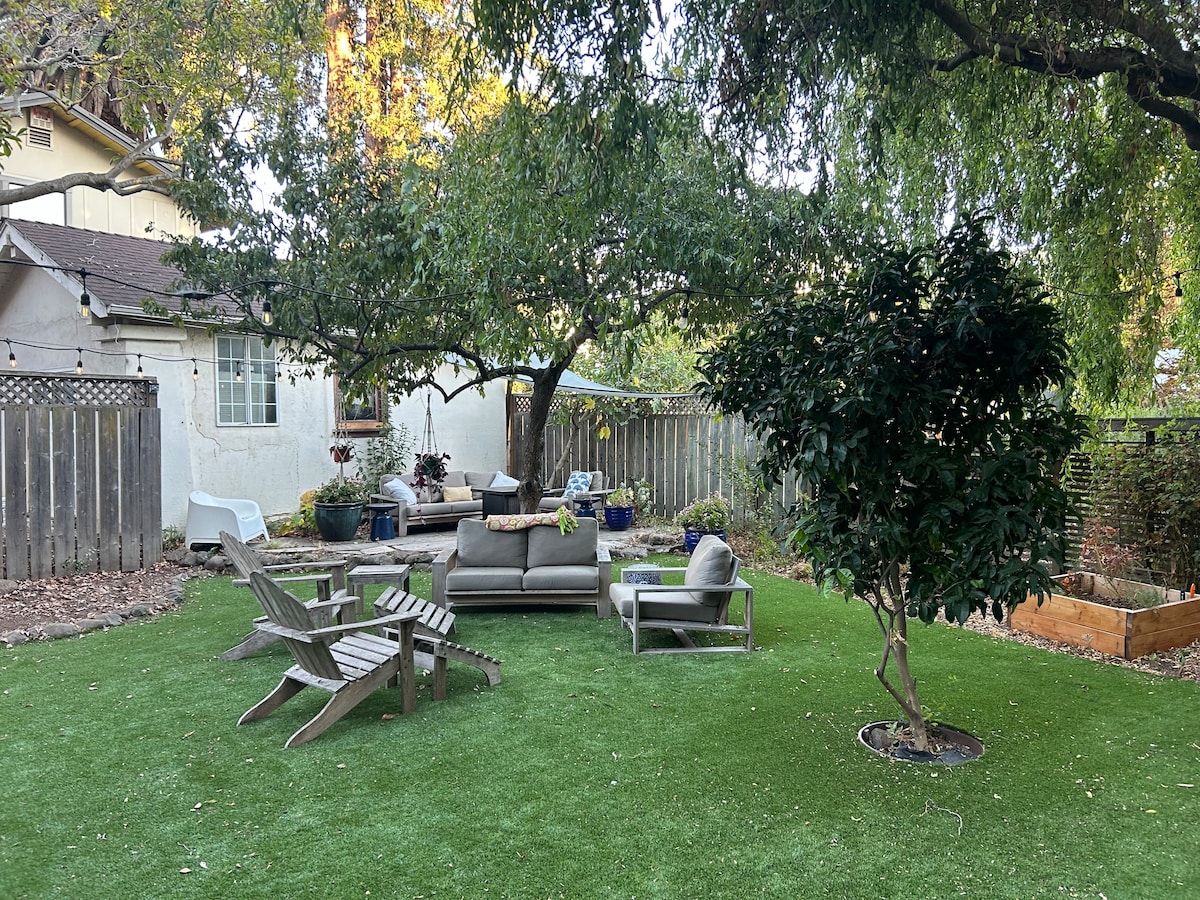 A spacious shared backyard is visible, featuring a lush green lawn and a large seating area with a comfortable couch and armchairs. Several potted plants add greenery, while a tree provides shade, creating a welcoming outdoor space for relaxation.