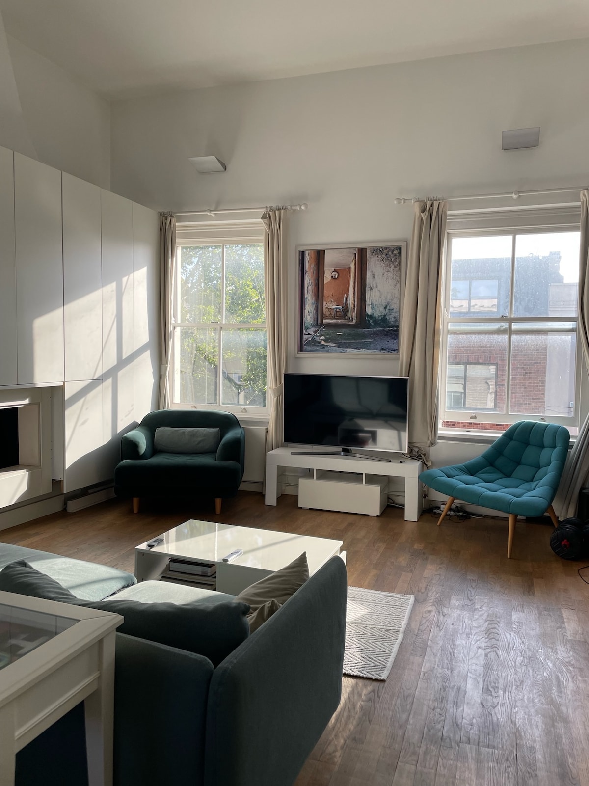 The living room features large windows allowing natural light to fill the space. Two seating areas are provided with a pair of armchairs, one in teal and one in a neutral color. A television is positioned on a light entertainment unit, with a small coffee table in front.