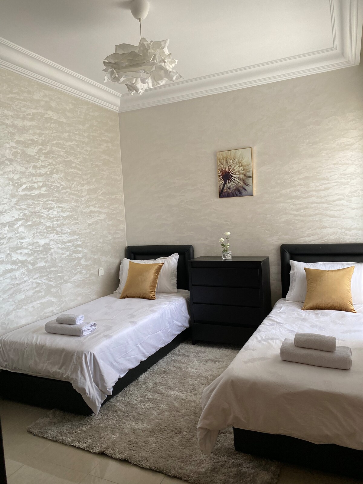 The room features two comfortable twin beds, each adorned with white linens and decorative pillows. A bedside table sits between the beds, accompanied by a small vase of flowers. The space is complemented by soft lighting and textured wall decor.