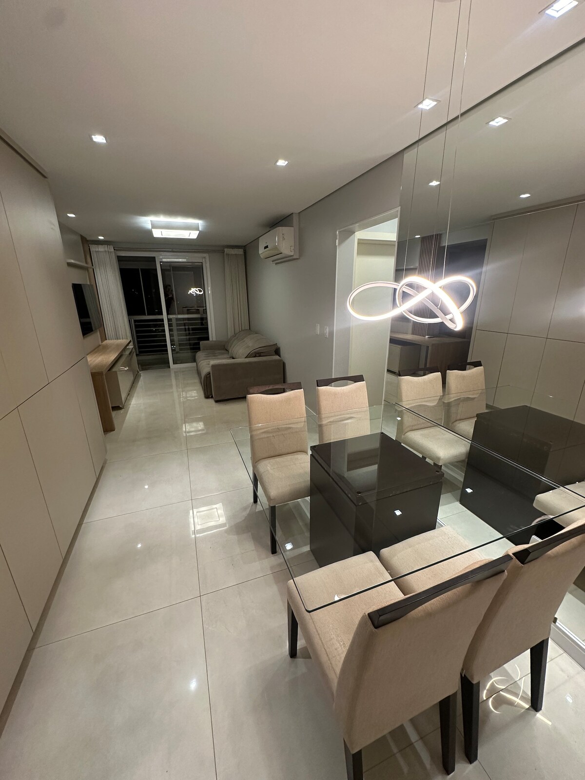 A modern dining area features a glass table surrounded by four upholstered chairs. A stylish light fixture hangs above, reflecting off the polished tile floor. A seating area is visible in the background, with sliding doors opening to a balcony.