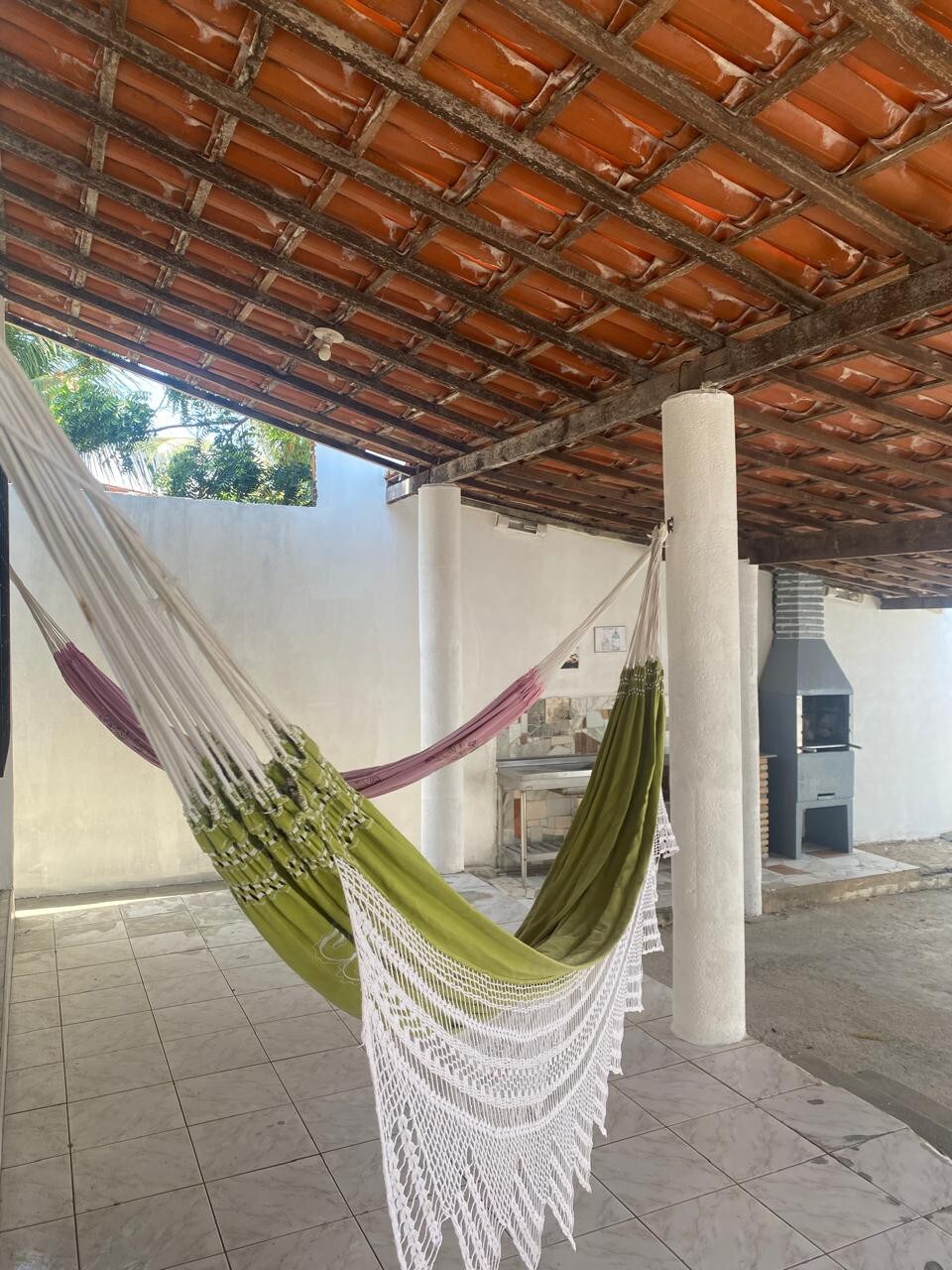 Two hammocks are suspended in a shaded outdoor area, underneath a textured red-tiled roof. A simple barbecue grill is positioned against one wall, and the floor is covered in light tiles. Natural light filters through the space, highlighting the serene environment.