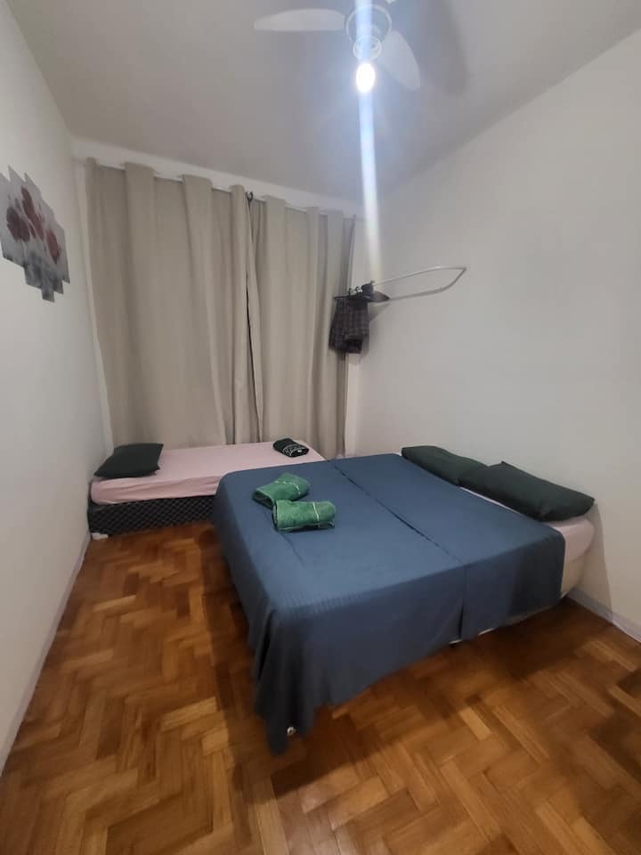 ห้องพักพร้อมเตียงเดี่ยวและเตียงคู่ เตียงพร้อมหมอนเตียงที่มีและไม่มียางยืดปลอกหมอนผ้าห่มและผ้าเช็ดตัว มีกระจกหน้าพัดลมเพดานมาคอว์พร้อมไม้แขวนเสื้อและม่านทึบแสงที่หน้าต่าง