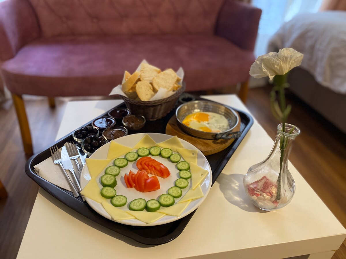 A breakfast tray is presented on a table, featuring a circular arrangement of cheese and tomato slices, complemented by cucumber slices. A bowl with bread and various small dishes including olives is included. A cooked egg is featured alongside a vase with a single flower.