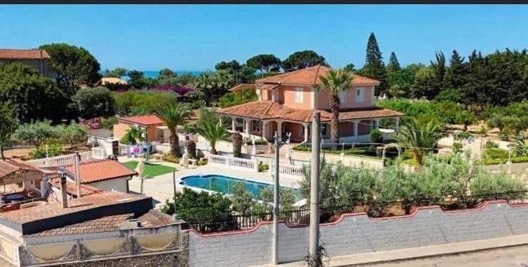 A spacious villa with a terracotta-tiled roof is surrounded by lush greenery, featuring palm trees and flowering plants. A large swimming pool is situated in the center of the garden. The distant view reveals the clear blue sea, enhancing the serene outdoor environment.