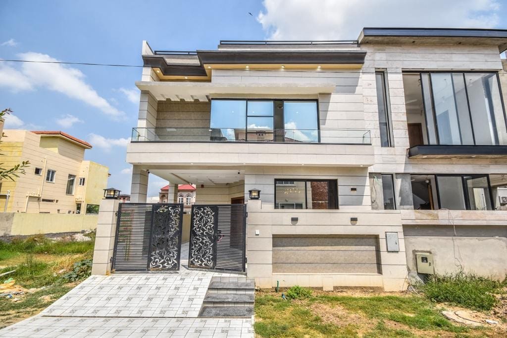 A modern two-story home features a sleek exterior with large glass windows. The entrance is framed by decorative gates, leading to a spacious patio area. Lush greenery surrounds the property, complemented by a bright blue sky above.