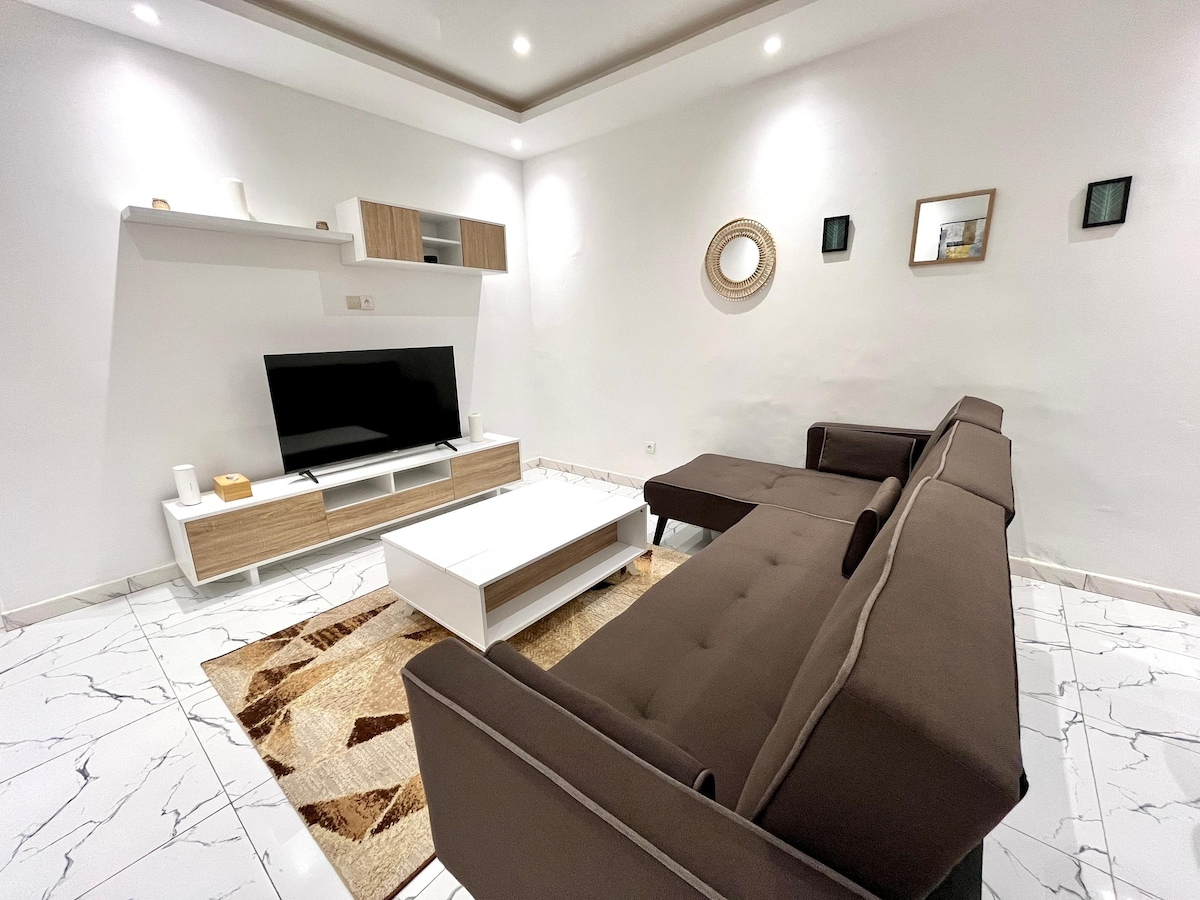 A modern living area features a sleek brown sofa with a chaise section, positioned adjacent to a white media console. A flat-screen television is displayed prominently on the wall. Decorative mirrors and small shelves add subtle character, while a patterned area rug enhances the room's contemporary feel.