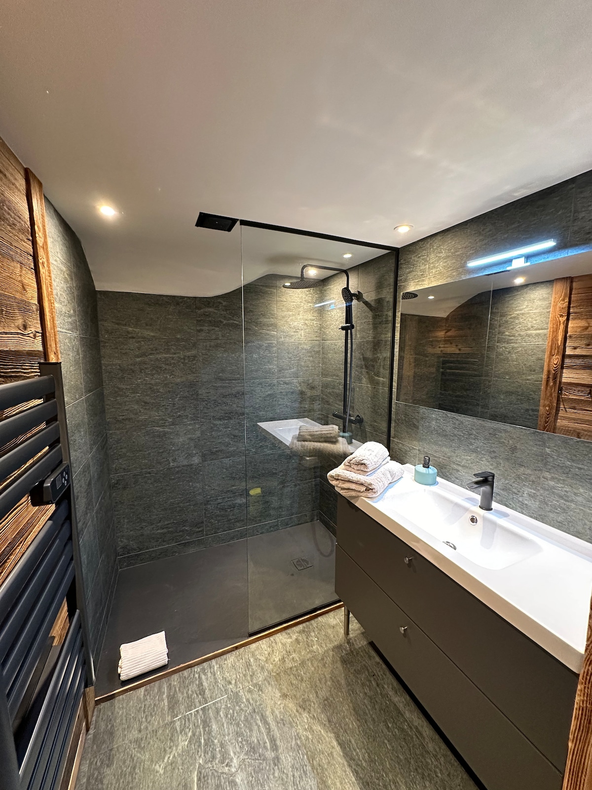 A modern bathroom features a glass shower enclosure with a rainfall showerhead. A sleek vanity unit with dual sinks is complemented by natural wood accents. Soft, folded towels are neatly arranged beside a small bottle of amenities, while dimmable lighting enhances the space.