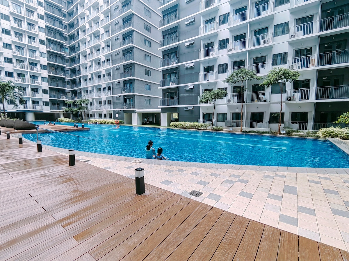 A swimming pool is highlighted at the center of the building complex, surrounded by stylish lounge chairs and a wooden deck. Residents can be seen enjoying the water. The surrounding area features greenery and modern architecture, creating a serene recreational space.