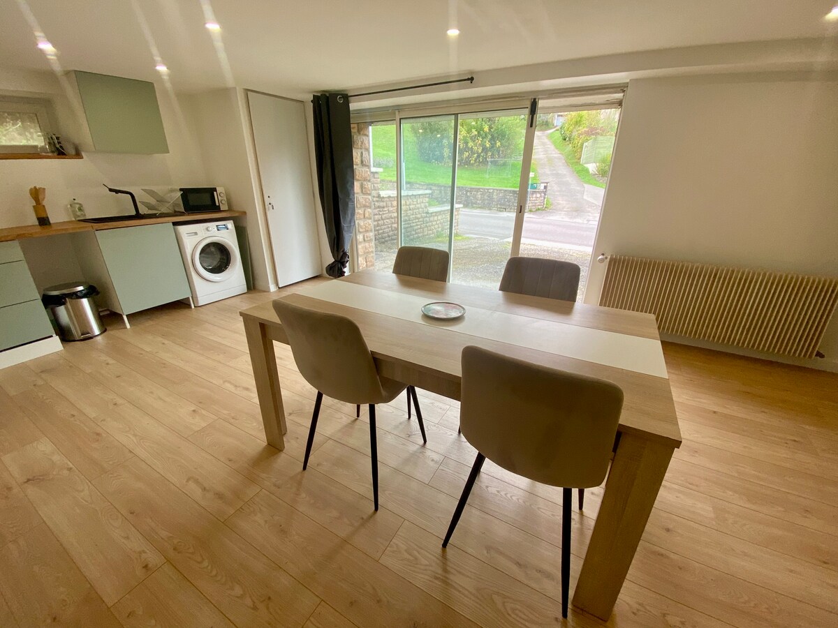 A bright dining area features a light wooden table accompanied by three upholstered chairs. Large glass doors allow natural light to fill the space, providing views of the surrounding outdoors. A washing machine and kitchen appliances are visible in the background.