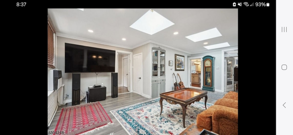 A spacious family room is captured, featuring a large flat-screen TV mounted on the wall. Natural light enters through the skylights above. A stylish coffee table sits on a decorative area rug, complemented by cozy seating options and a wooden cabinet. Two guitars are displayed nearby.