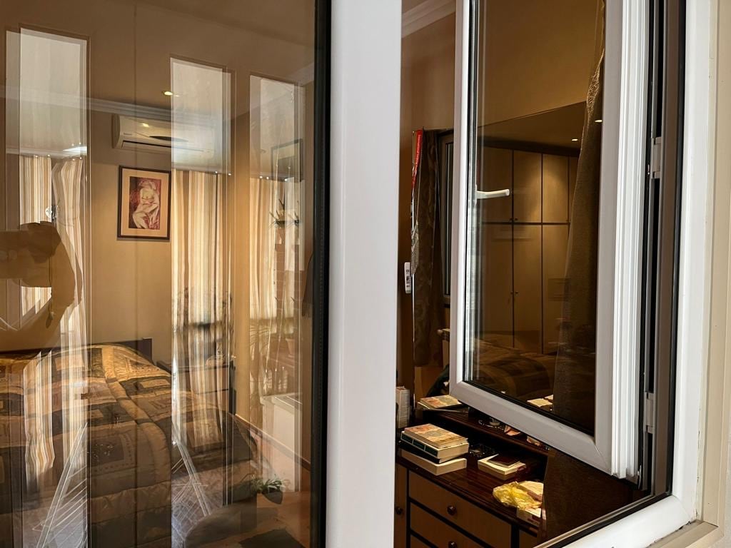 A glimpse into a well-lit bedroom through a window, featuring warm light filtering through sheer curtains. A spacious bed is visible, with decorative artwork adorning the walls and a dresser displaying books and personal items. Air conditioning is mounted above for comfort.