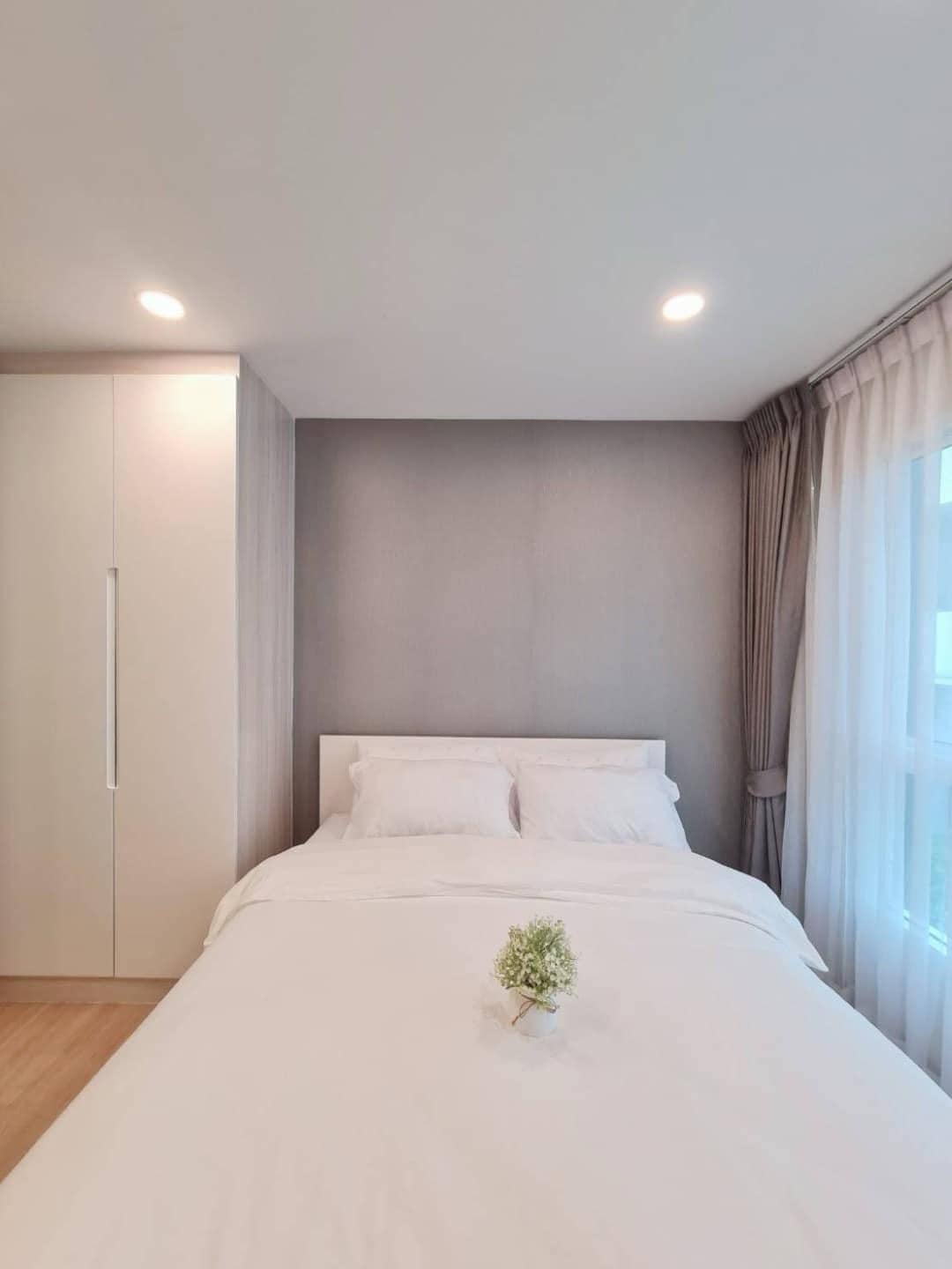 A light and airy bedroom features a spacious bed dressed in crisp white linens. Soft, natural light filters through sheer curtains, illuminating the subtle grey accent wall. A small plant decorates the center of the bed, adding a touch of greenery to the space.