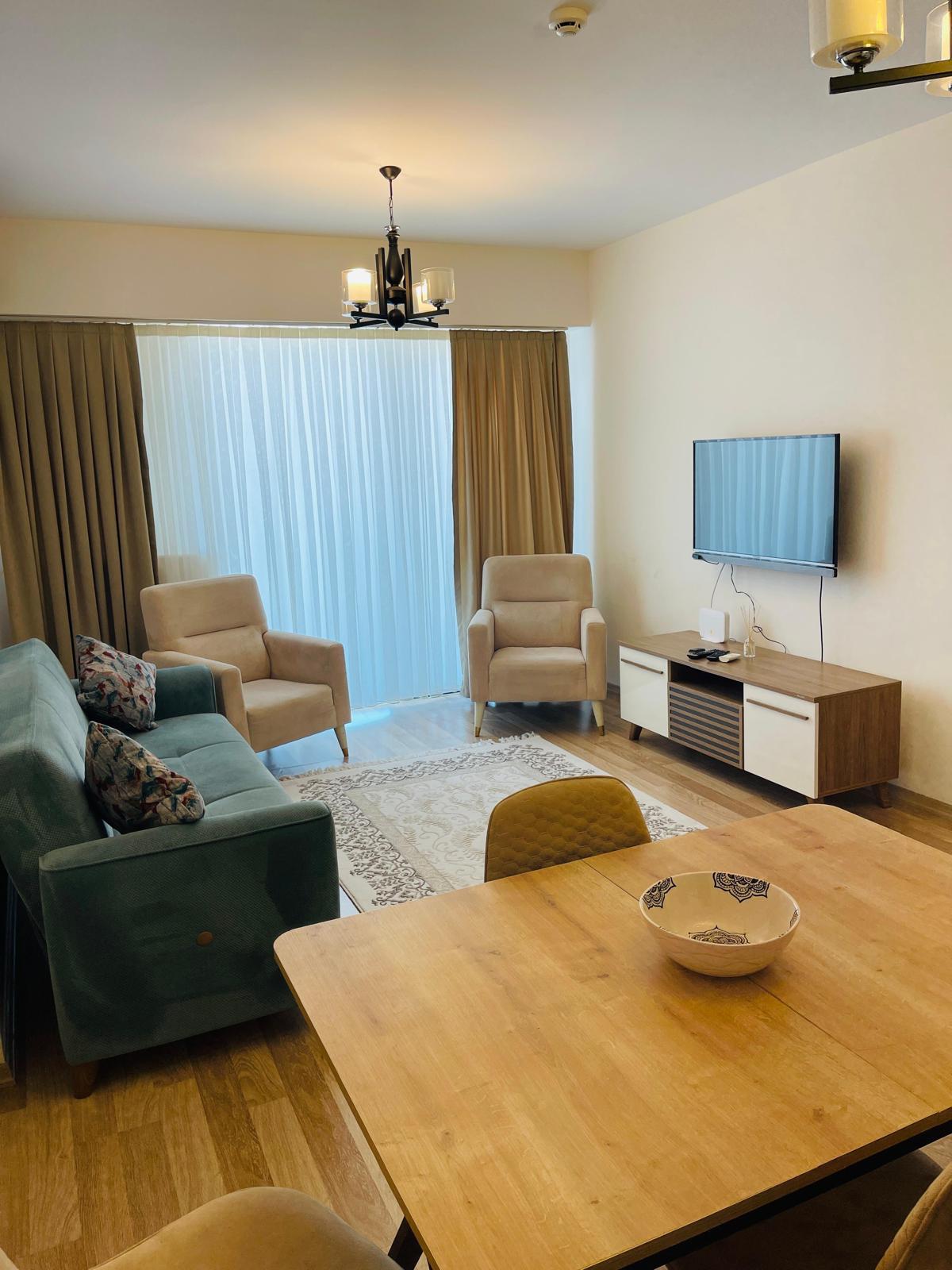 A cozy living area features a teal sofa and two beige armchairs, complemented by a wooden coffee table. A television is mounted on the wall opposite a window with sheer curtains that allow natural light to filter in, enhancing the inviting space.