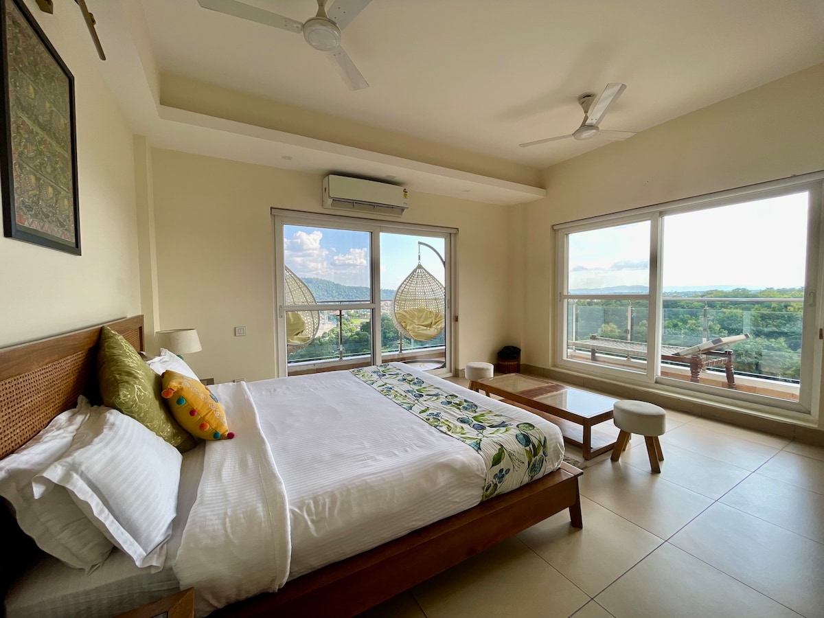 A spacious bedroom features a large bed with decorative pillows and crisp linens. Large windows offer expansive views of surrounding greenery and distant hills. Two hanging chairs add a unique touch, while a small wooden table and stool enhance the comfortable ambiance.