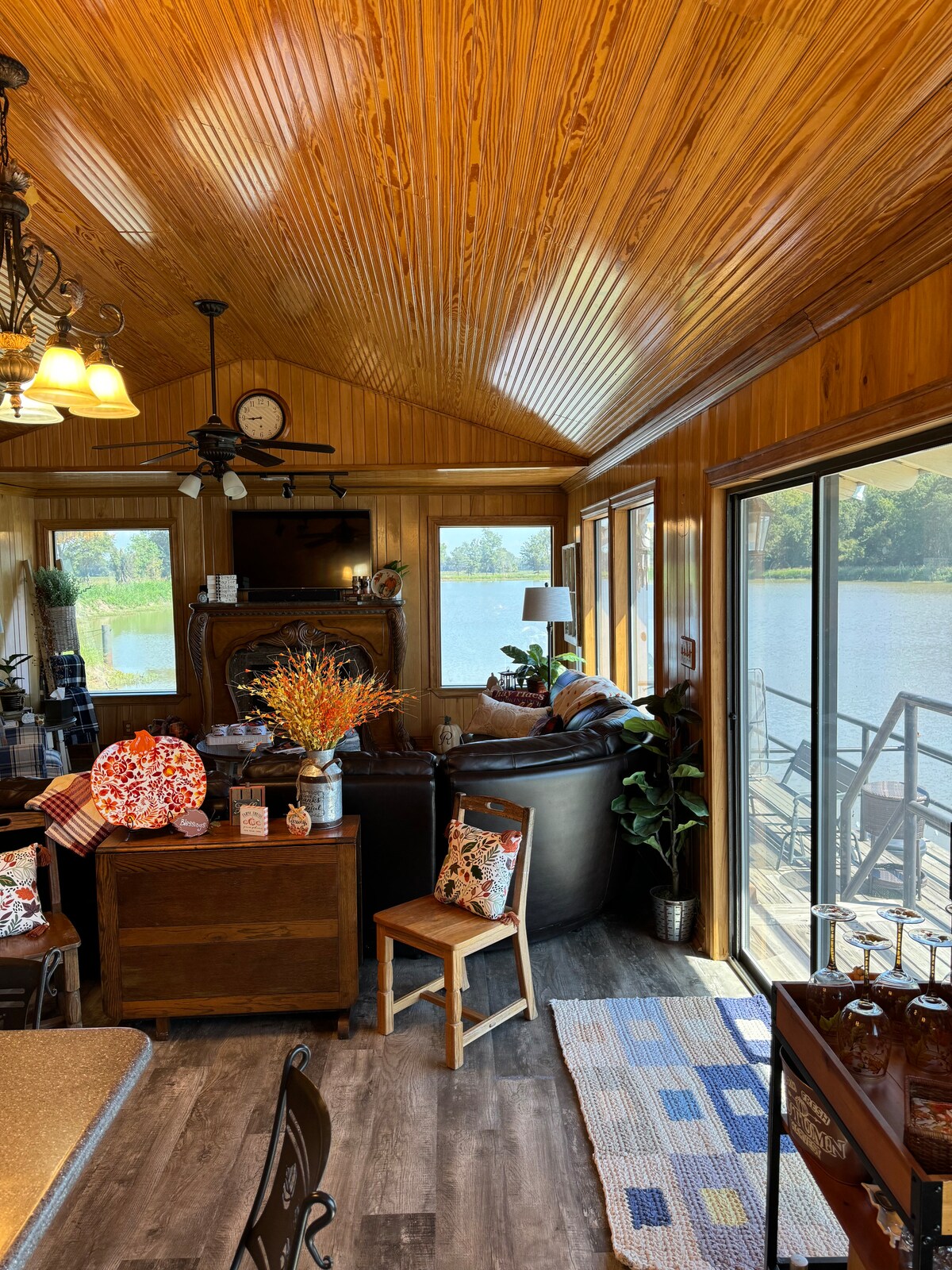 The interior space features wooden paneling and a high ceiling, creating a warm atmosphere. Large windows provide views of the water, while a cozy seating area includes a dark leather sofa and decorative accents. A dining area is visible with a wooden table and chairs.