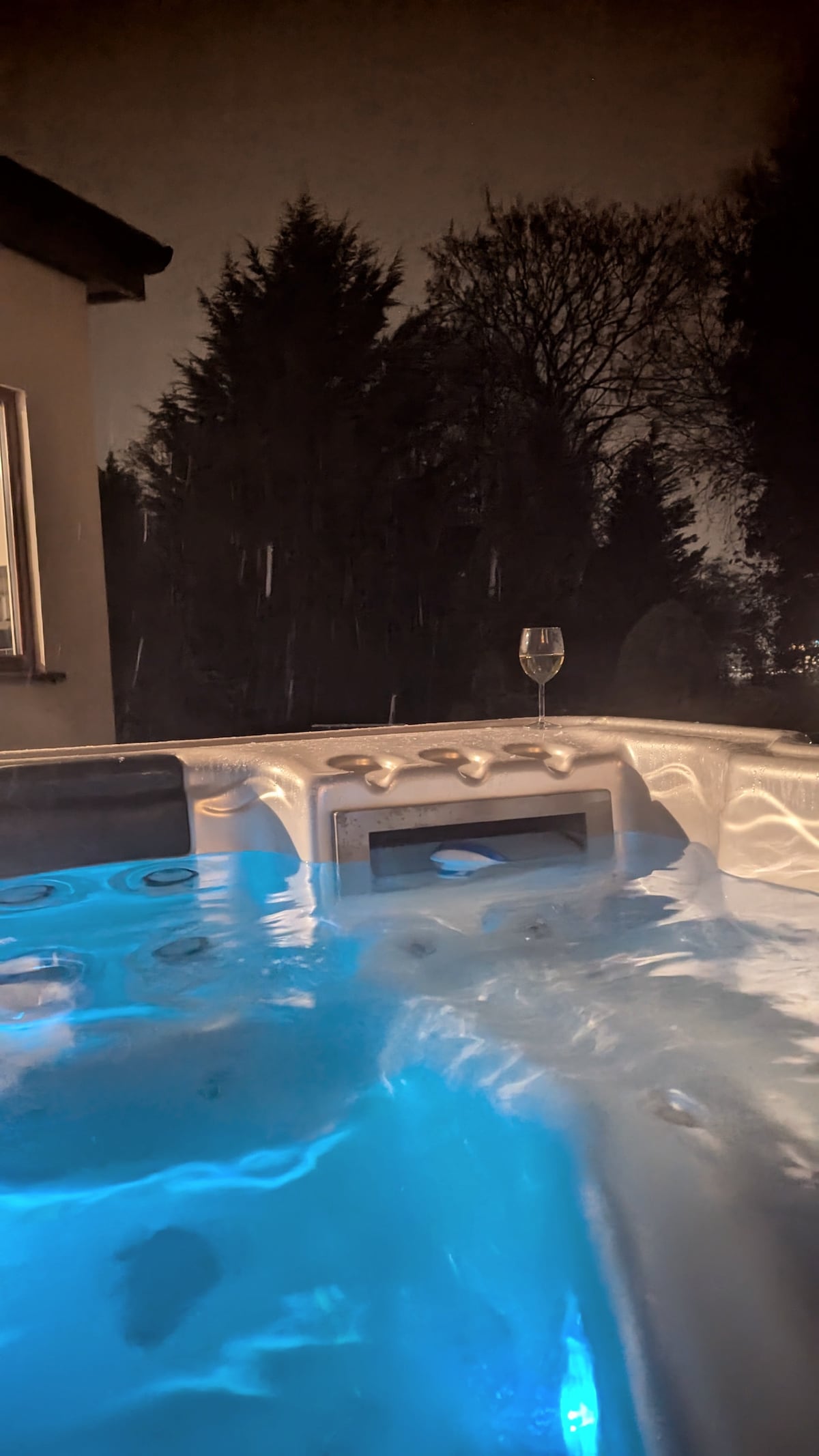 A hot tub is seen at night, with gentle blue lighting illuminating the water. A glass rests on the edge, surrounded by dark trees in the background and a window reflecting soft light from the house.