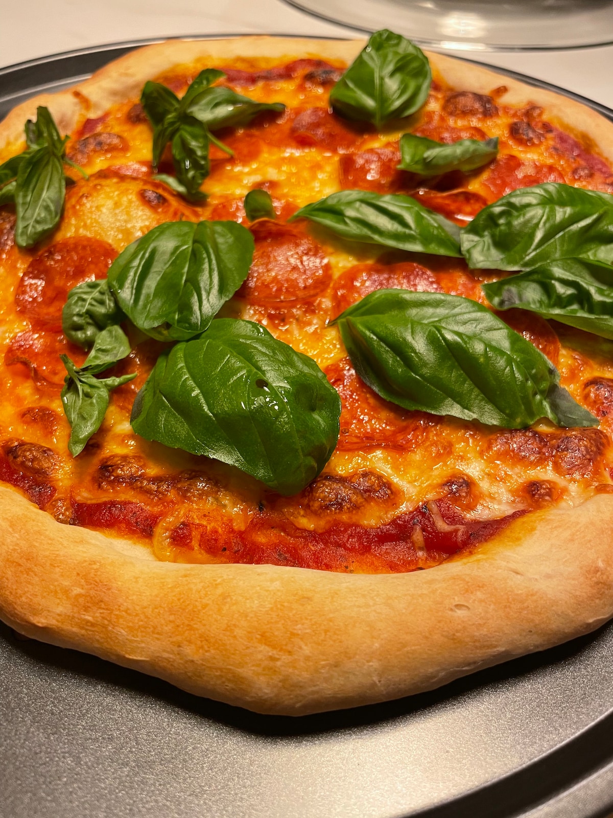 A freshly baked pizza is placed on a round black tray. The golden crust surrounds a layer of melted cheese, sliced pepperoni, and vibrant green basil leaves, adding a pop of color to the dish. The pizza appears inviting and warm.