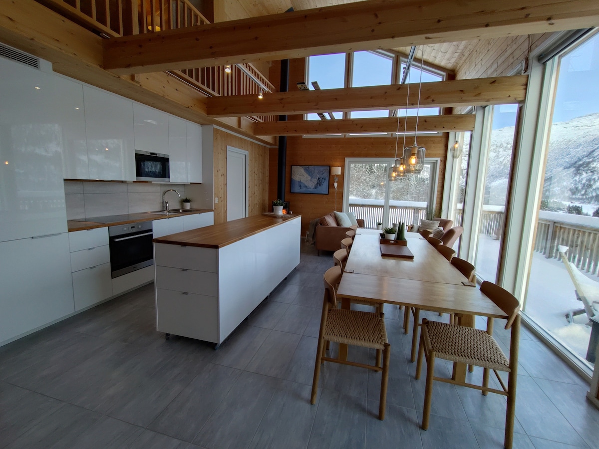 A spacious kitchen and dining area features modern cabinetry and a large wooden table with several chairs. Large windows allow natural light to illuminate the room, offering views of the snow-covered landscape outside. An island with counter space provides additional work area.