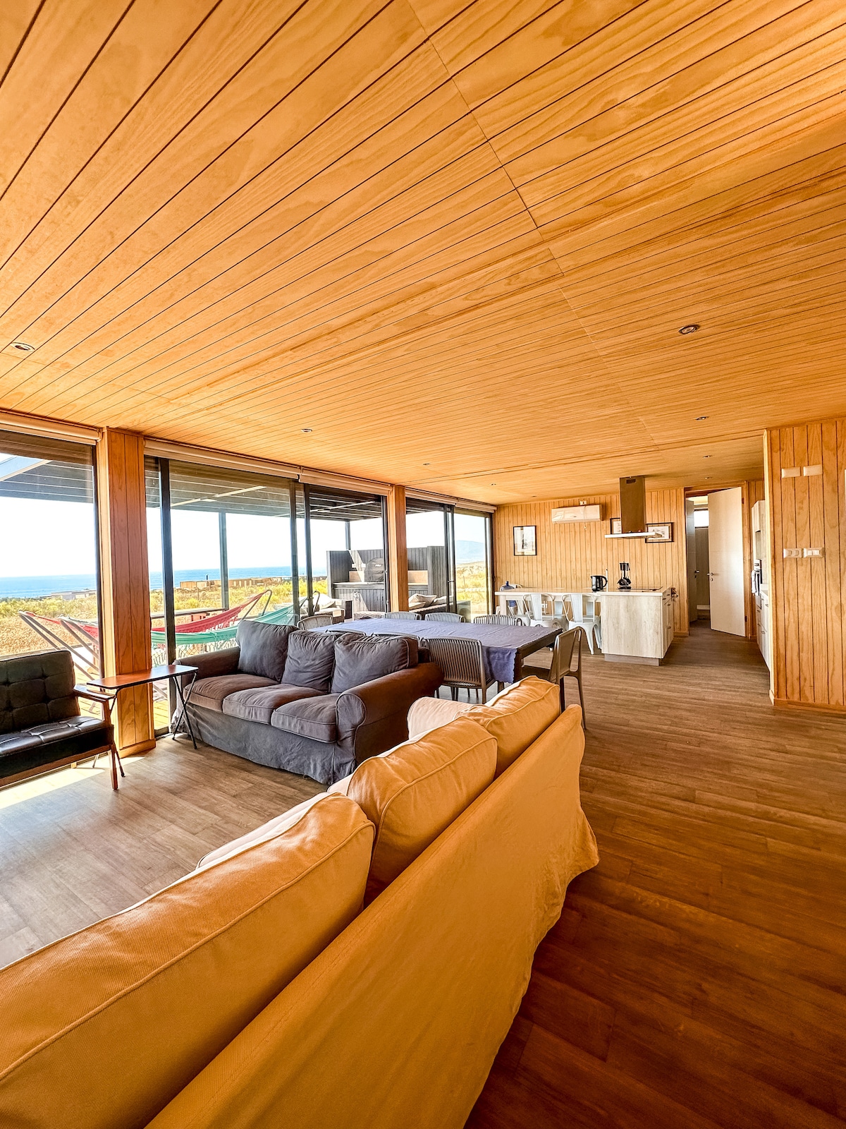 A spacious living area features warm wooden paneling and large glass windows offering views of the surrounding landscape. Comfortable seating is arranged around a light-hued sofa and armchairs. The kitchen area, partially visible, includes modern appliances and is seamlessly integrated into the open layout.